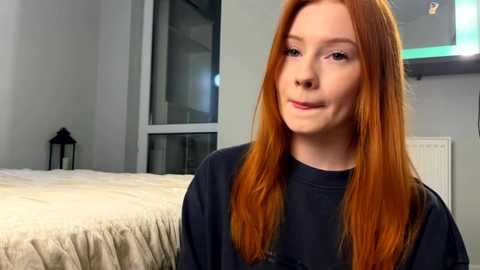 Video of a fair-skinned, red-haired young woman with straight hair, wearing a black sweater, standing in a modern, minimalist bedroom with a white bed and gray walls.