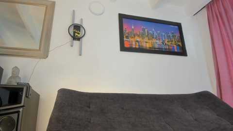 Video of a minimalist, modern bedroom with a dark gray sofa, a colorful cityscape painting, a framed mirror, and a wall-mounted clock on a white wall.