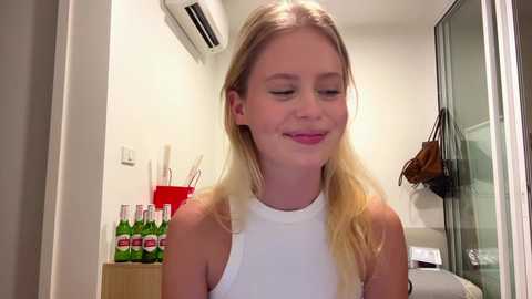 Video of a smiling young blonde woman in a white sleeveless top, standing in a modern, minimalistic room with a glass shower door and a red bucket filled with green beer bottles.
