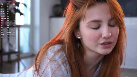 Video of a young, fair-skinned woman with long, red hair and a small nose ring, wearing a white, floral-patterned shirt, eyes closed in a serene expression, sitting indoors with a blurred background.