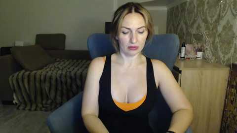 Video of a fair-skinned, blonde woman in a black tank top with orange lining, sitting in a blue armchair. Background includes a dark couch, patterned wallpaper, and a wooden side table with bottles.