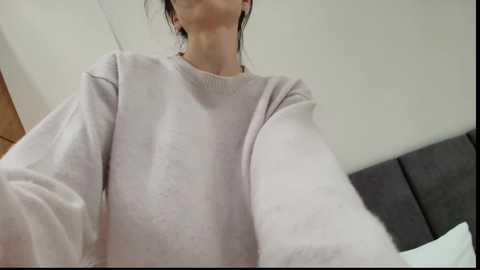 A video of a woman in a light gray, oversized sweater, her dark hair pulled back, standing in a room with white walls and a gray upholstered headboard.