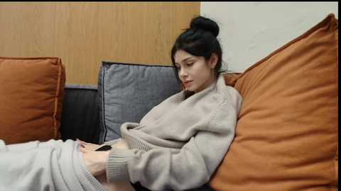 Video of a young woman with dark hair in a bun, wearing a beige sweater, sitting on a sofa with orange and gray cushions, reading a book.