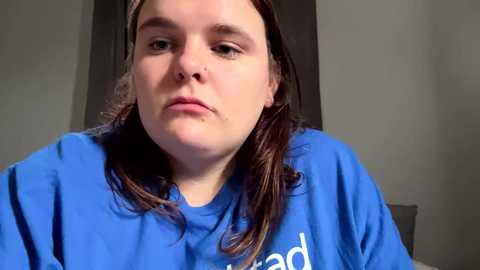 Media: Video of a plus-size woman with long brown hair, wearing a blue shirt, looking serious in a neutral-toned room with a dark doorway.