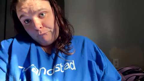 Video of a woman with shoulder-length brown hair wearing a blue \"Lulastar\" t-shirt, making a pouty face, indoors against a gray wall, with a purple bag in the background.