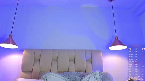 Video of a modern bedroom with a beige, tufted headboard, two hanging pendant lights, a gray bedspread, and a digital clock displaying 4:23 on the right wall.