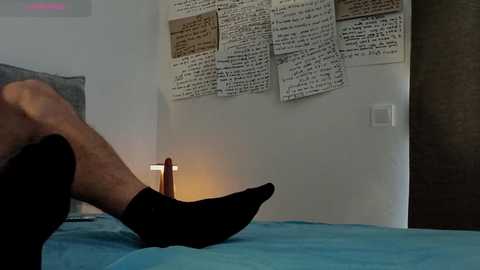 A video shows a person in black socks sitting on a blue bed, with a lamp and white wall with pinned papers in the background.