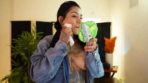 Video of a woman with long dark hair, wearing a denim jacket, applying makeup with a green brush, tattoos on her chest, in a bright, modern room with plants and a lamp.