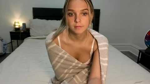 Video of a blonde woman in a plaid blanket, sitting on a bed with white sheets, in a dimly lit, minimalist bedroom.