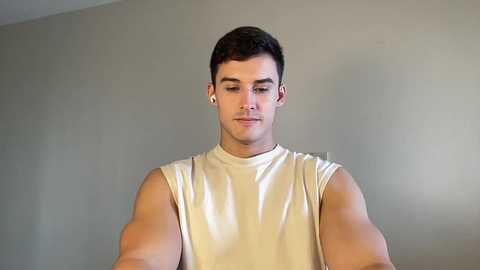 Video of a young man with short dark hair, wearing a sleeveless white tank top, standing against a plain, light gray wall. He has a neutral expression.