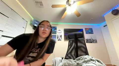 Video of a young woman with long brown hair and glasses, wearing a black T-shirt, sitting on a black leather gaming chair in a modern, brightly lit bedroom with neon lights, posters, and a ceiling fan.
