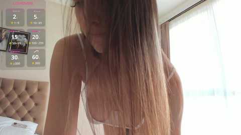 A video of a slender, light-skinned woman with long, straight brown hair, wearing a white lace bra, leaning forward, partially visible in a bright, modern bedroom.
