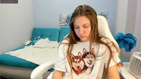 Video of a teenage girl with long brown hair, wearing a white T-shirt with a graphic of two huskies, sitting on a white chair in a hospital room with turquoise bedding and blue wall art.