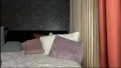 Video of a neatly made bed with white linens, a pink pillow, and a mauve quilted blanket against a dark, patterned wall. A tall, cream-colored curtain and a vibrant orange curtain frame the bed.
