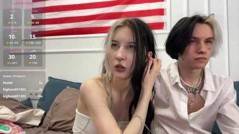Video of a young woman with long blonde hair and a young man with short black hair, both wearing casual clothing, seated on a couch. An American flag hangs in the background.