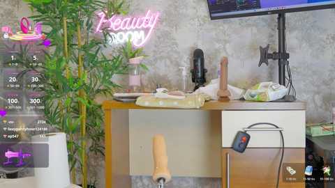 A video of a cluttered room with a beige desk, a laptop, a dildo, and a sex toy stand, with a neon \"Beauty\" sign and a bamboo plant in the background.