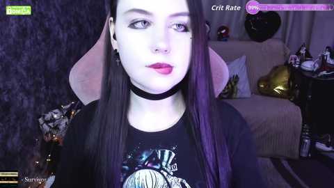 A video of a pale-skinned woman with long black hair, wearing a black choker and a graphic t-shirt, in a dimly lit room with dark walls and a plush couch.