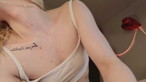 Video of a woman with fair skin, wearing a beige tank top, displaying a small tattoo of Chinese characters on her upper back. Background features a red heart-shaped lamp and neutral-toned wall.