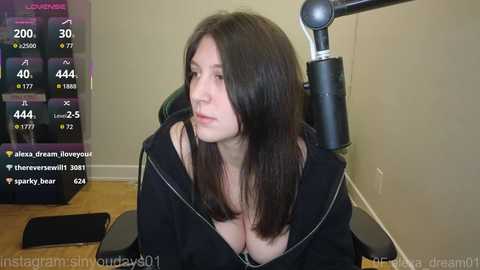 Video of a young woman with long dark hair, wearing a black hoodie, sitting on a black chair in a dimly lit room with a monitor displaying streaming statistics.