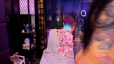 Video of a modern, dimly lit room with purple and blue lighting. A tattooed woman in a white lace dress stands near a glass window with a cityscape view. A pink garment hangs on a chair.