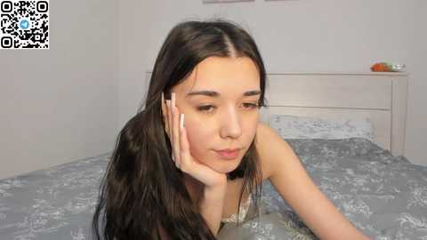 Video of a young woman with long black hair, pale skin, and a slender physique, lying on a bed with a floral-patterned duvet, resting her head on her hand, in a minimalist bedroom setting.