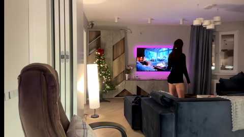 Video of a modern living room with a woman in a black dress playing video games, a lit Christmas tree, and a TV screen displaying a game.