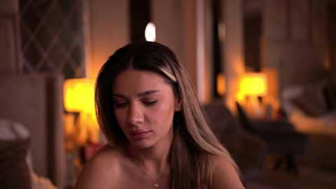 Video of a young woman with light brown skin and long, straight, blonde hair, wearing a headband, sitting indoors in a dimly lit room with warm, ambient lighting, looking down thoughtfully.