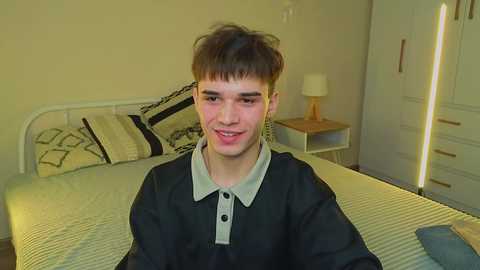 Video of a young man with short brown hair, wearing a black polo shirt, smiling in a sparsely decorated bedroom with a white bed and minimalist furniture.