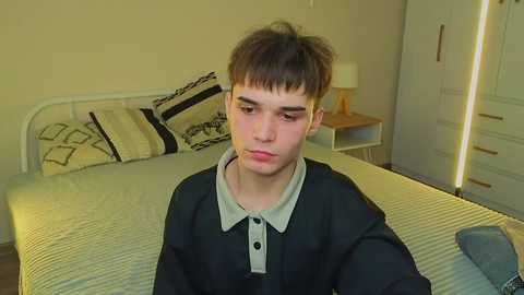 A video of a young man with straight, dark hair, wearing a black polo shirt with a gray collar, sitting on a bed with beige bedding and patterned pillows in a simple, beige-walled room.