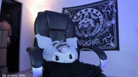 Video of a black leather office chair with a plush white bunny wearing a black hat and scarf, in a room with a black and white tapestry featuring a skull and crossed bones, under dim blue lighting.