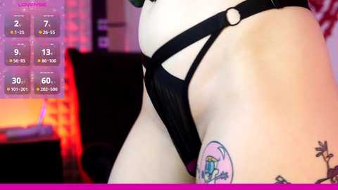 Video of a fair-skinned woman in black strappy lingerie, with visible tattoos, including a colorful anime character, in a dimly lit room with a computer screen displaying a game interface.