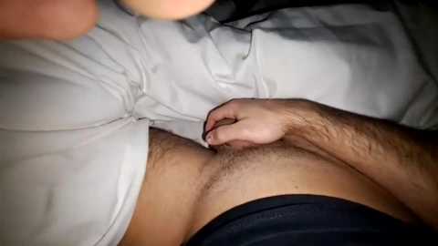 A close-up video of a man's stomach with a light tan, hairy chest, and a hand adjusting dark underwear, lying on a bed.