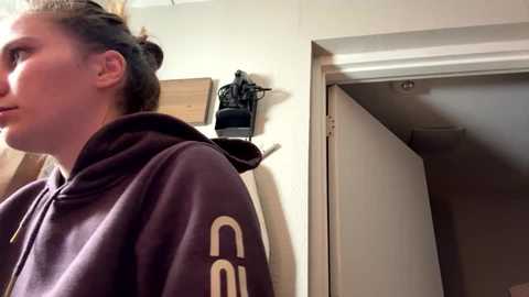 Video of a young woman with light skin and dark hair tied back, wearing a dark purple hoodie with white \"UNO\" text on the sleeve, standing in a doorway.