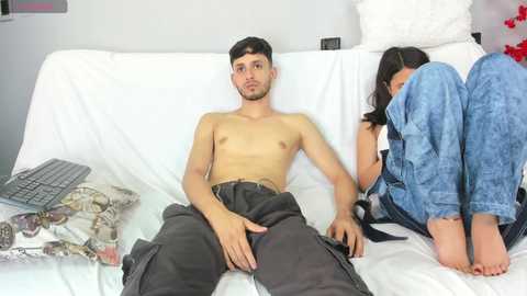 Video of a shirtless young man with a slender build and dark hair, sitting on a white couch. A woman in blue jeans is asleep beside him, with a blanket covering her legs.