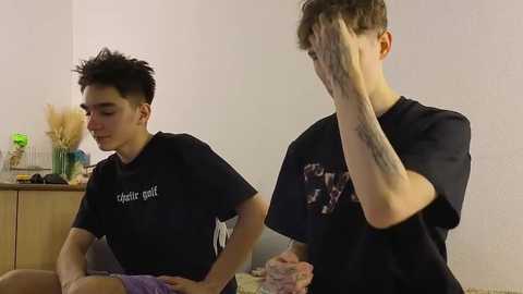 Video of two young men in casual black T-shirts; one covers face, the other looks down. Background features a wooden dresser with decorative items and a beige wall.