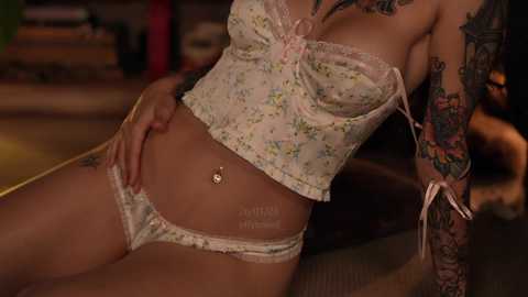 Video of a tattooed woman in a floral lingerie set, showcasing her pierced navel, sitting on a couch in a dimly lit room.