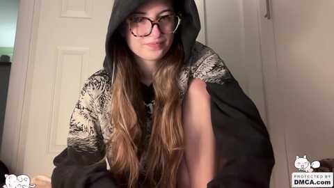 Video of a young woman with long, wavy brown hair, wearing a black hooded cloak with a white pattern, round glasses, and a confident expression, indoors near a white door.