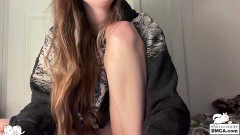 Video of a young woman with long, wavy brown hair, wearing a black and white leopard-print hoodie, sitting indoors with a white door and part of a bed visible in the background.