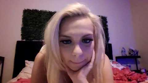 A video of a blonde woman with fair skin and a slender build, lying on a bed with red and pink bedding, resting her head on her hand. The room features a dark headboard, a green wall hanging, and a nightstand with items.