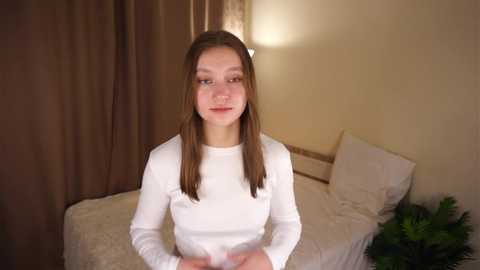 Video of a young Caucasian girl with light skin and straight brown hair, wearing a white long-sleeve shirt, sitting on a bed with white bedding and pillows.