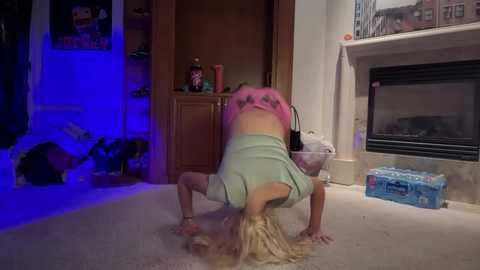 Media: Video of a young woman with blonde hair in a green top performing a handstand in a living room with a blue light, wooden shelves, a fireplace, and a water bottle on the carpet.