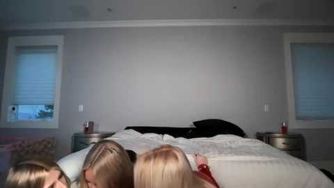 Video of a modern bedroom with a gray wall, two white window blinds, a black headboard, and three people sleeping on a white bed with white pillows.