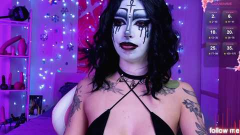 Video of a person with pale skin and dark, wavy hair, wearing a black halter top, heavy makeup, tattoos, and a choker, in a dimly lit room with purple and blue lights, and a calendar displaying 26th April.