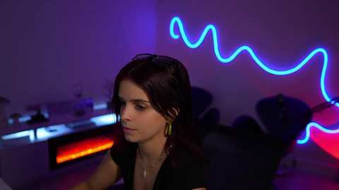 Video of a young woman with dark hair, wearing headphones, seated in a dimly lit room with neon blue and red lighting, a modern desk, and electronic devices.