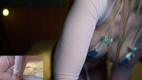 Video of a blonde woman in a white, long-sleeved top, revealing her cleavage, in a dimly lit room with a wooden wall and a bed with patterned bedding.