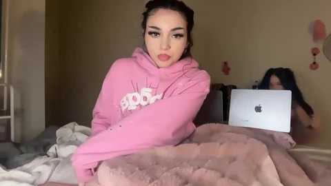 Video of a young woman with olive skin, dark hair, and pink hoodie, sitting on a bed with a laptop open, blurred background of a room with pink walls and posters.