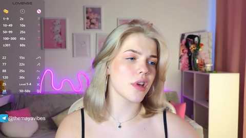 A video of a young, fair-skinned, blonde woman with shoulder-length hair, wearing a black top, standing indoors with a pink neon light background, framed by various floral and decorative items.