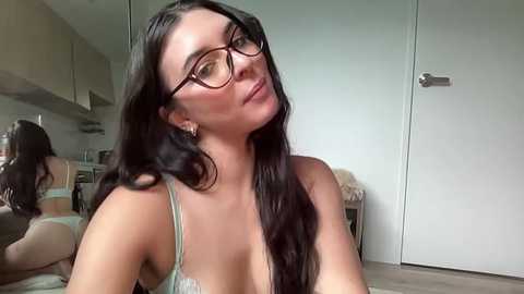 A video of a young woman with long black hair, wearing glasses and a light green lace bra, sitting in a modern kitchen with white cabinets and a white door in the background.