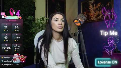 Video of a woman with long black hair, wearing a white top, sitting in a room with a TV displaying \"Tip Me\" and \"Loveense ON\" icons.