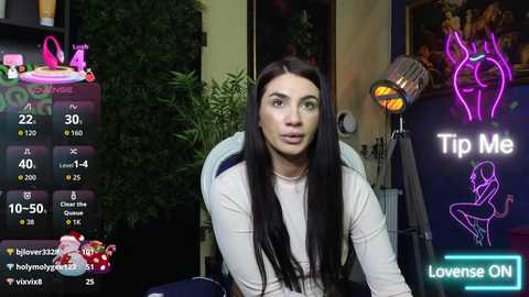 A video of a woman with long black hair, wearing a white top, sitting on a chair in a room with neon signs and a green plant in the background.